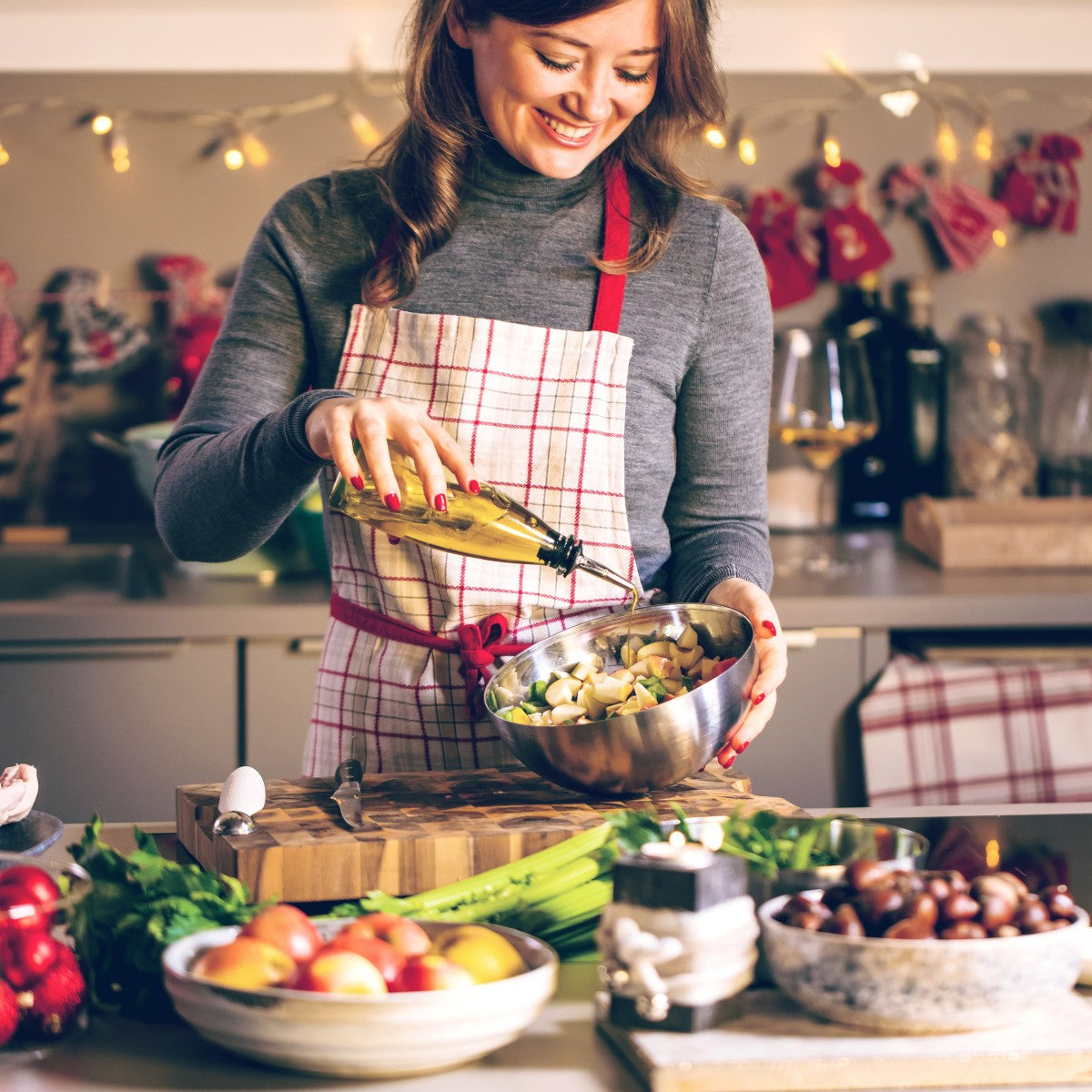 A woman in a gray sweater and plaid apron smiles as she drizzles olive oil into a bowl of chopped vegetables while preparing food in a festive kitchen decorated with lights.