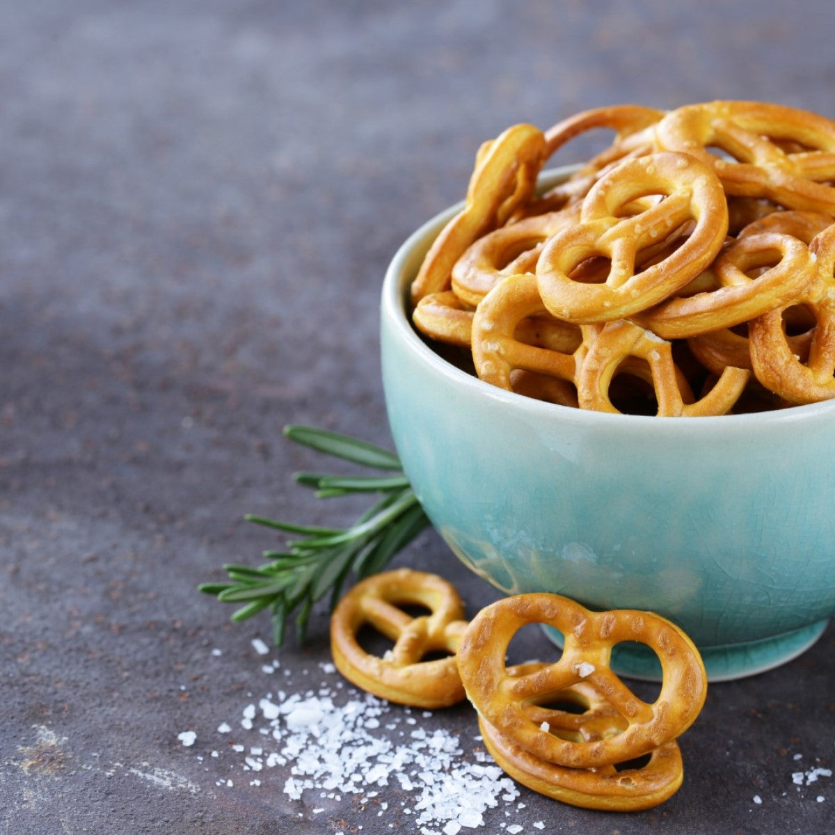 A turquoise bowl filled with crunchy pretzels sits on a textured surface, accompanied by a sprig of rosemary and a sprinkle of coarse salt, highlighting a savory snack option.