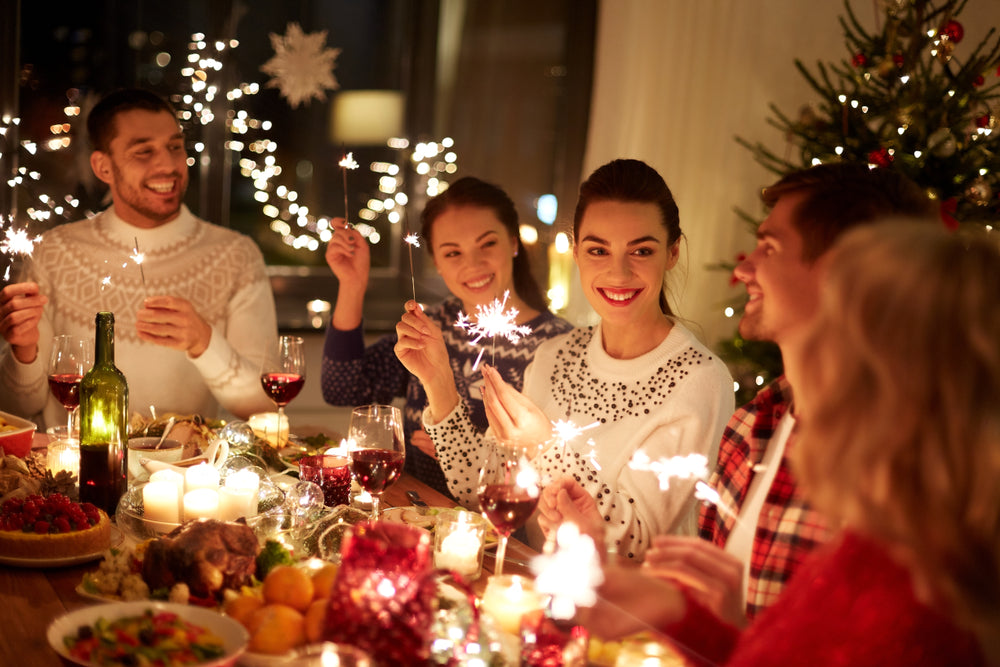 A group of five friends joyfully celebrate at a festive dinner table, holding sparklers, surrounded by holiday decorations, food, and candles, with a Christmas tree in the background.