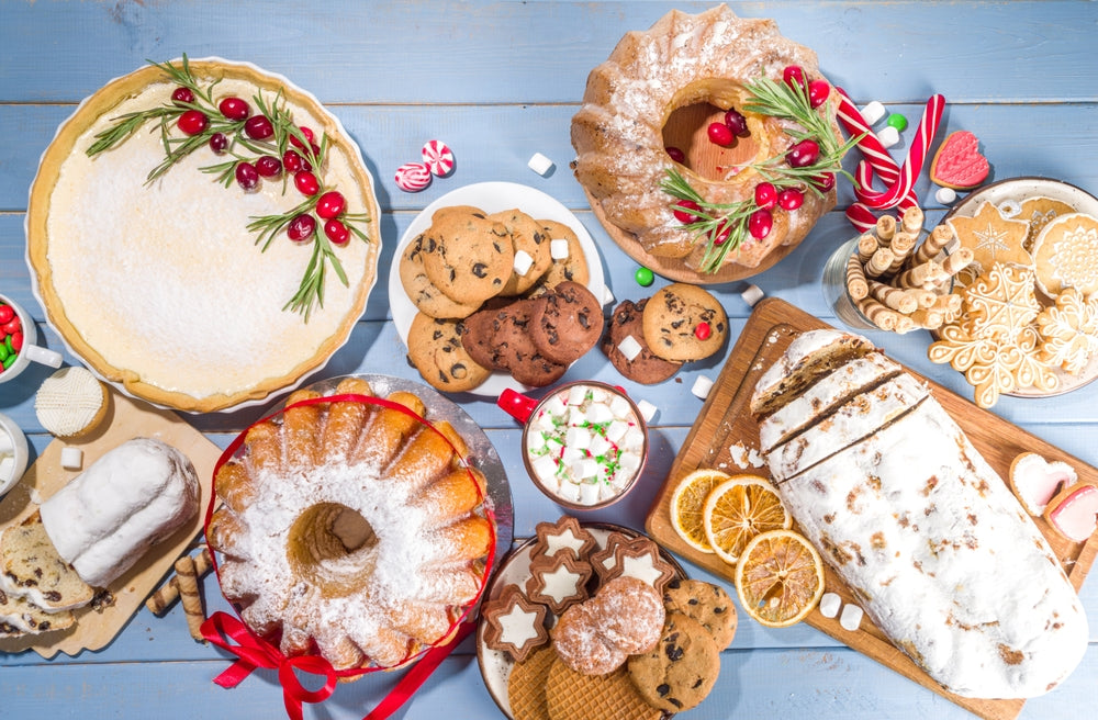 A festive spread of bariatric-friendly desserts includes a pie topped with cranberries, a bundt cake, cookies, gingerbread, and a loaf, all arranged on a light blue wooden table.