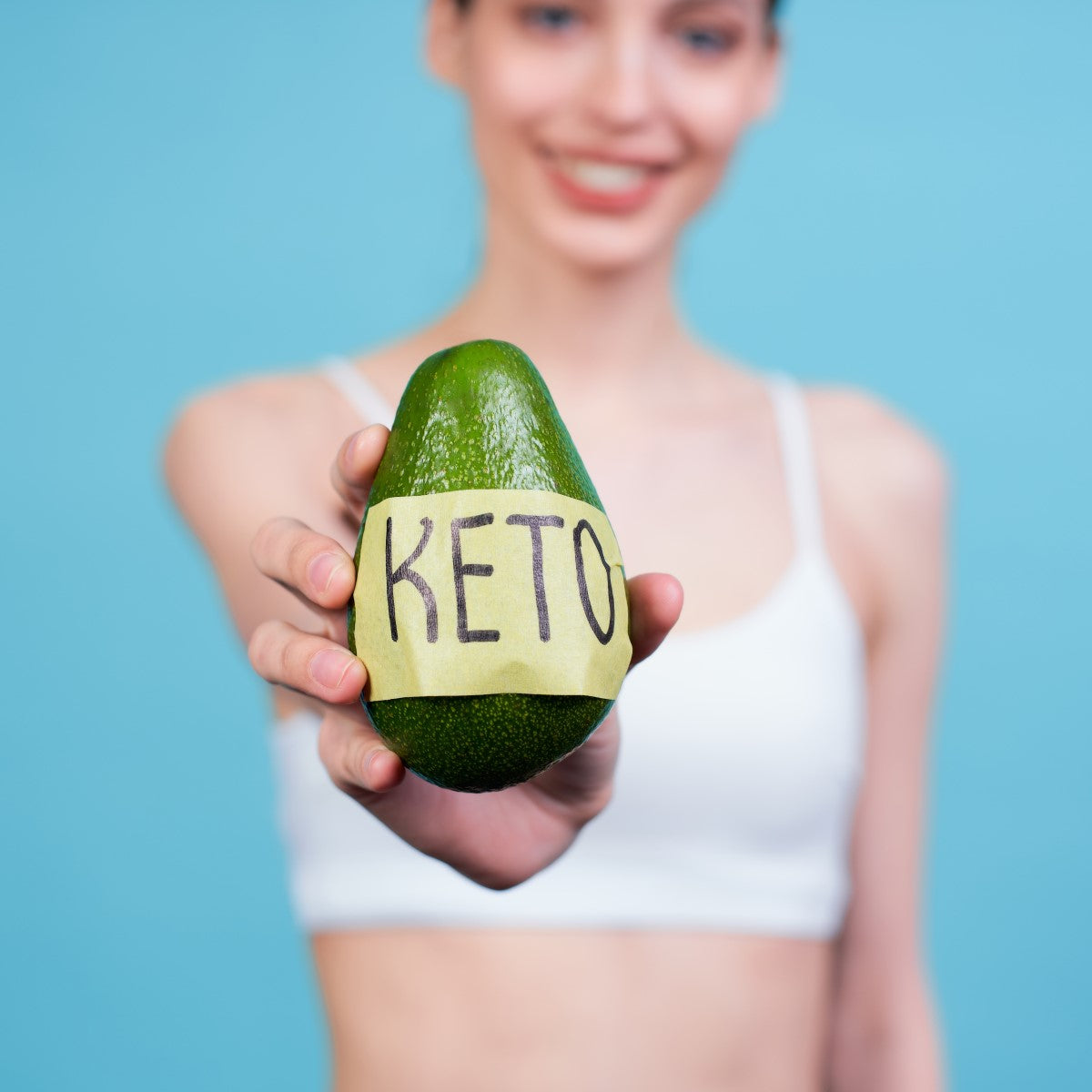 A smiling woman in a white tank top holds out an avocado with a yellow label that says "KETO," against a light blue background.
