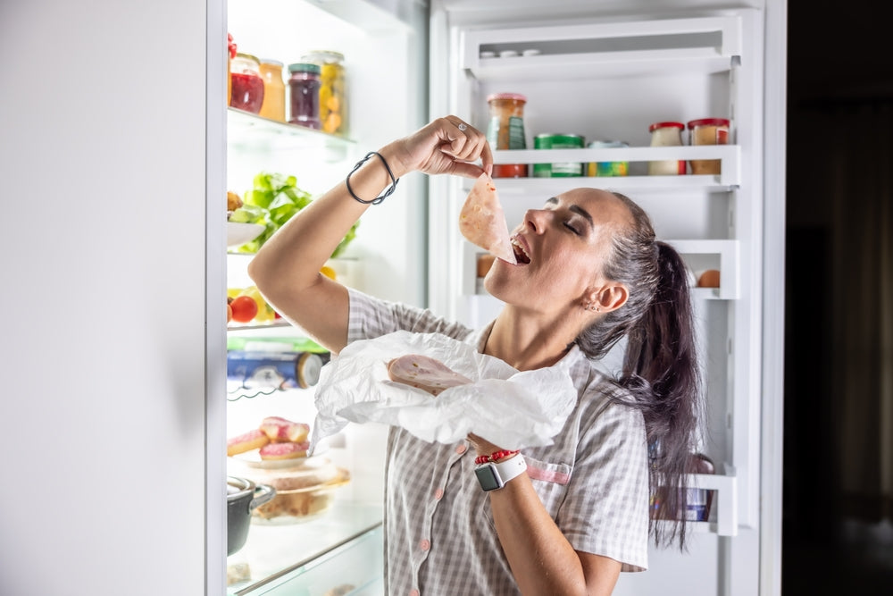 A woman in a kitchen enjoys a slice of turkey while standing in front of an open refrigerator filled with healthy food options, showcasing strategies for satisfying late-night cravings.