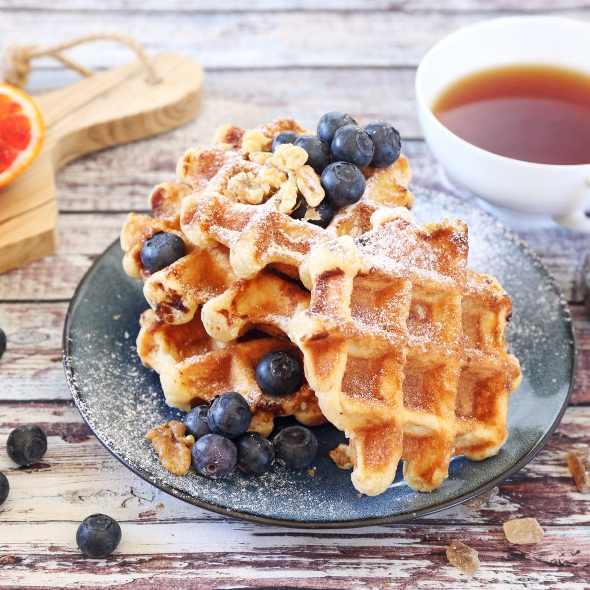 A stack of golden-brown waffles topped with blueberries and walnuts sits on a blue plate, dusted with powdered sugar, alongside a cup of tea and a wooden cutting board with a slice of orange.