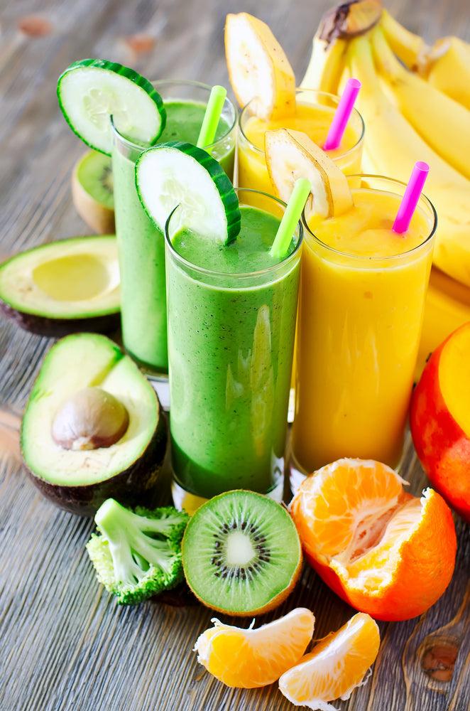 Four colorful smoothies in tall glasses, two green and two yellow, are displayed on a wooden table surrounded by fresh fruits like bananas, oranges, kiwi, and avocado, highlighting a nutritious high-protein diet.