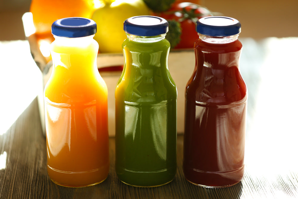 Three colorful juice bottles—orange, green, and red—are arranged on a wooden surface, with a blurred background of fresh fruits and vegetables, suggesting healthy liquid options for post-surgery recovery.