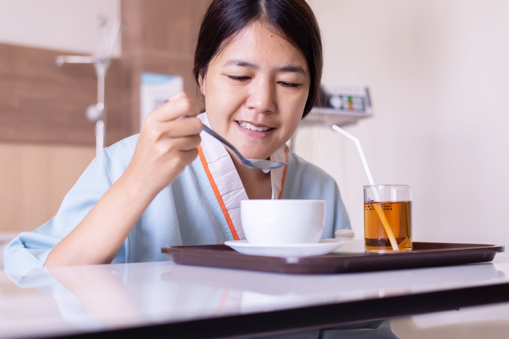 A woman in a hospital gown smiles as she eats soup with a spoon and drinks tea from a glass with a straw, sitting at a tray in a medical setting.