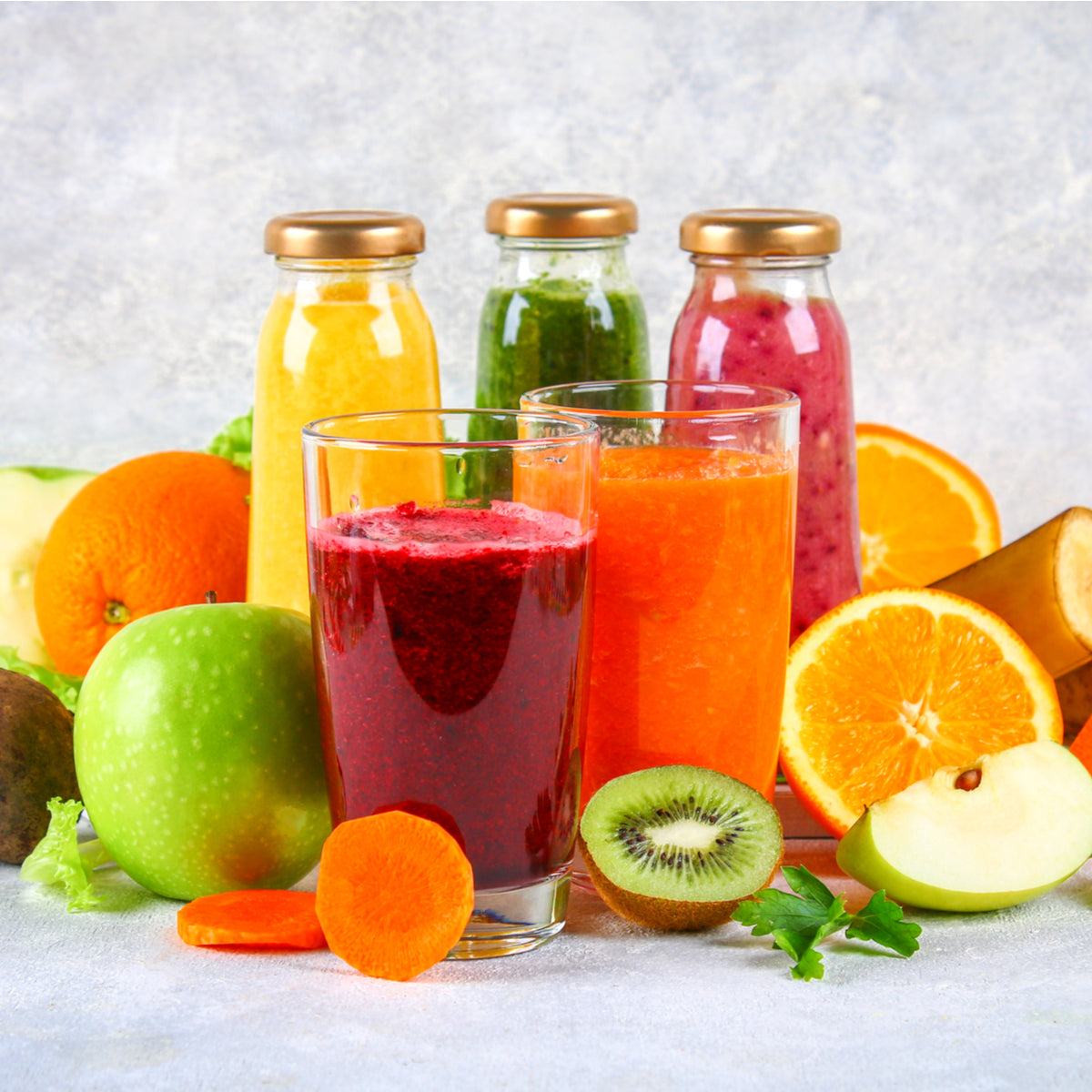 Two glasses of colorful smoothies sit in front of various fresh fruits and vegetables, with several bottles of juice in the background, all arranged on a light surface.