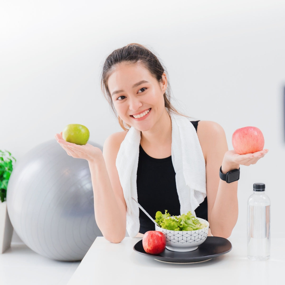 A smiling woman holds a green apple in one hand and a red apple in the other, with a bowl of salad and a water bottle on the table, set in a bright, modern kitchen environment.