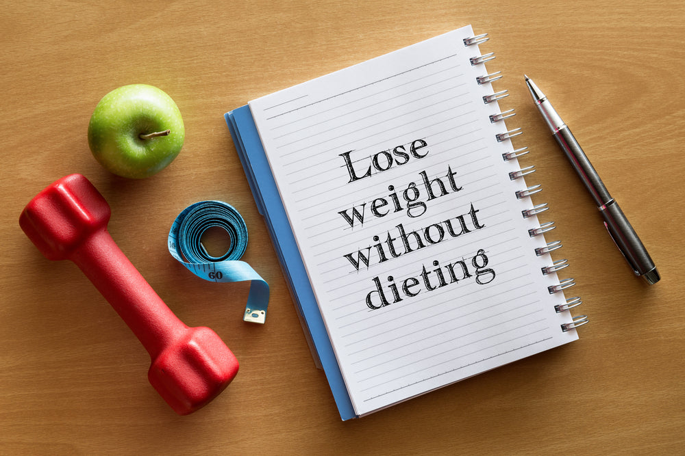 A red dumbbell, a green apple, a measuring tape, and a notebook with "Lose weight without dieting" written on the page are arranged on a wooden surface alongside a pen.