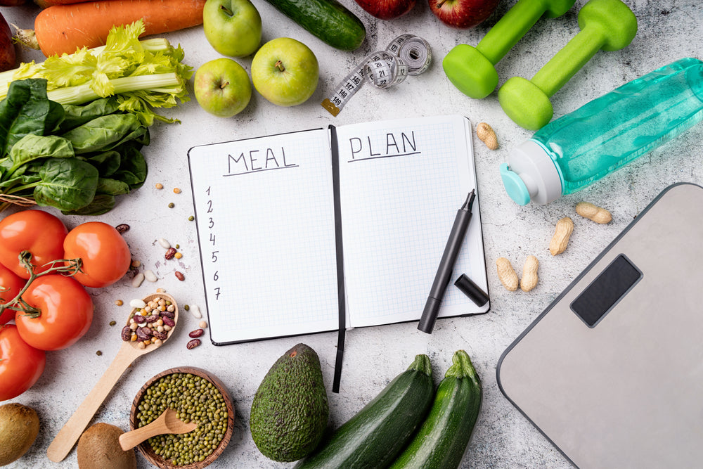 A notebook with "MEAL PLAN" written on it sits among fresh vegetables, fruits, a water bottle, a scale, and workout equipment, suggesting a focus on healthy meal planning and fitness.
