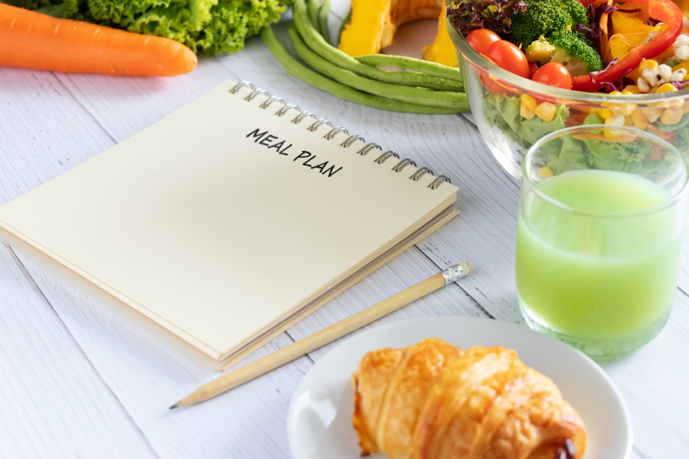A notepad labeled "MEAL PLAN" sits next to a plate with a croissant, a glass of green juice, and a colorful salad bowl, surrounded by fresh vegetables like carrots and lettuce on a wooden surface.