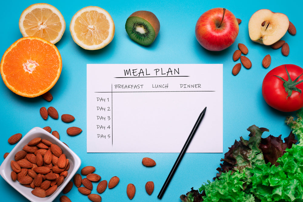 A blank meal plan sheet labeled "MEAL PLAN" is surrounded by fresh fruits like oranges, apples, and kiwis, along with nuts and leafy greens, set against a bright blue background.