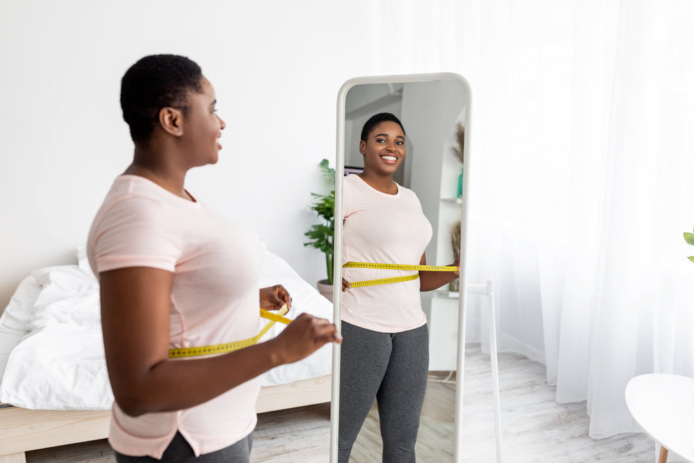 A smiling woman measures her waist with a tape measure while looking at her reflection in a mirror, standing in a bright, airy bedroom with a bed and plants in the background.