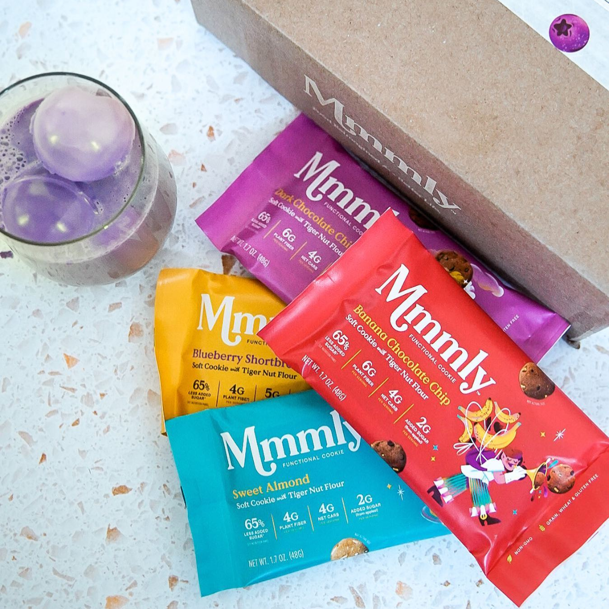 A variety of Mmmly Low-Carb Soft Cookie packages in vibrant colors are arranged on a textured surface, alongside a glass of purple drink with ice, showcasing flavors like Blueberry Shortbread and Banana Chocolate Chip.