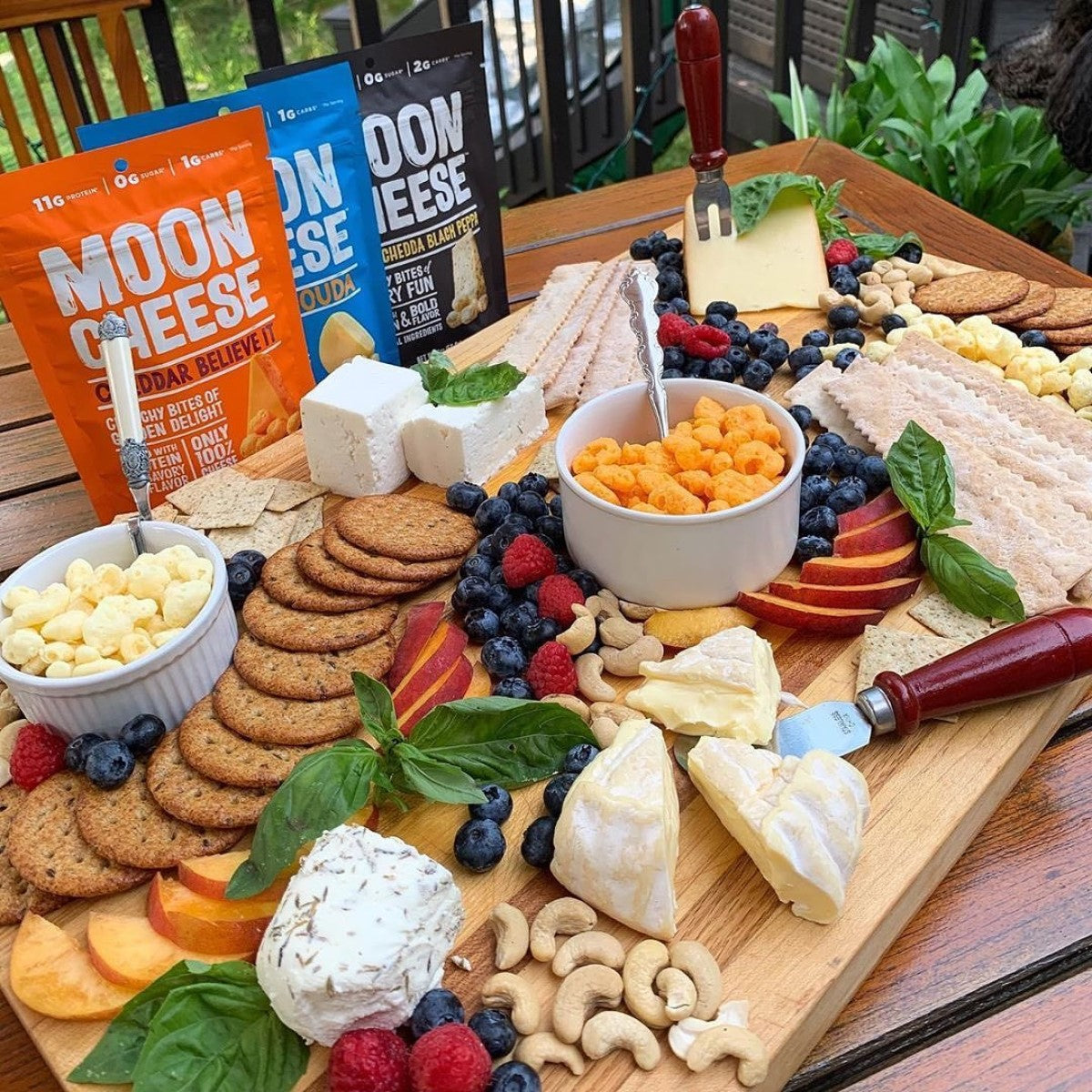 A wooden platter displays an assortment of Moon Cheese snacks, crackers, fresh fruits, and nuts, with vibrant colors and textures, set outdoors on a table surrounded by greenery.