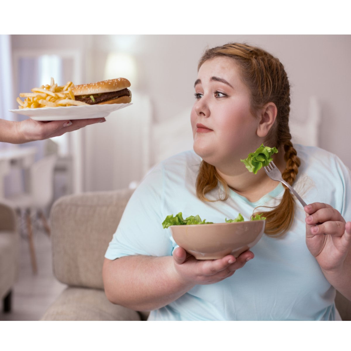A woman sitting on a couch holds a bowl of salad in one hand and a fork in the other, looking at a plate of fries and a burger being offered to her by another person.