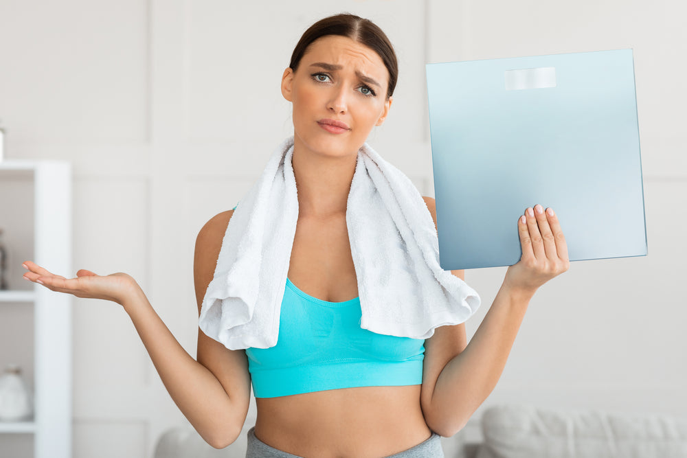 A woman in a turquoise sports bra and towel looks confused while holding a scale, standing in a bright room with light-colored walls, reflecting her frustration about losing inches but not weight.
