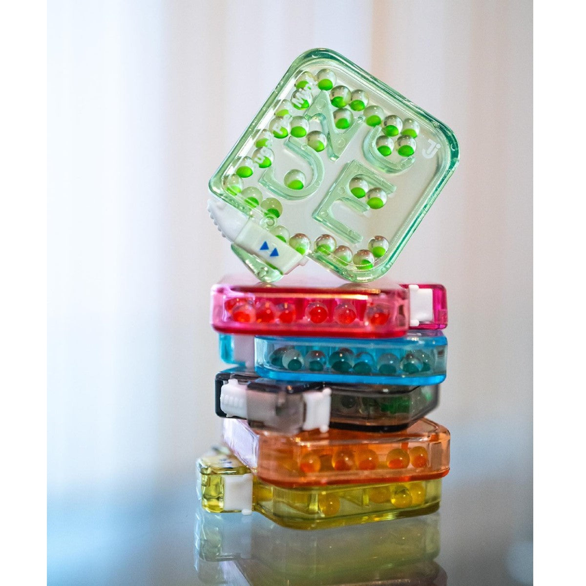 A stack of colorful NUDE Breath & Gut Freshener packs, featuring green, pink, blue, and orange containers, is displayed on a reflective surface with soft, natural light in the background.