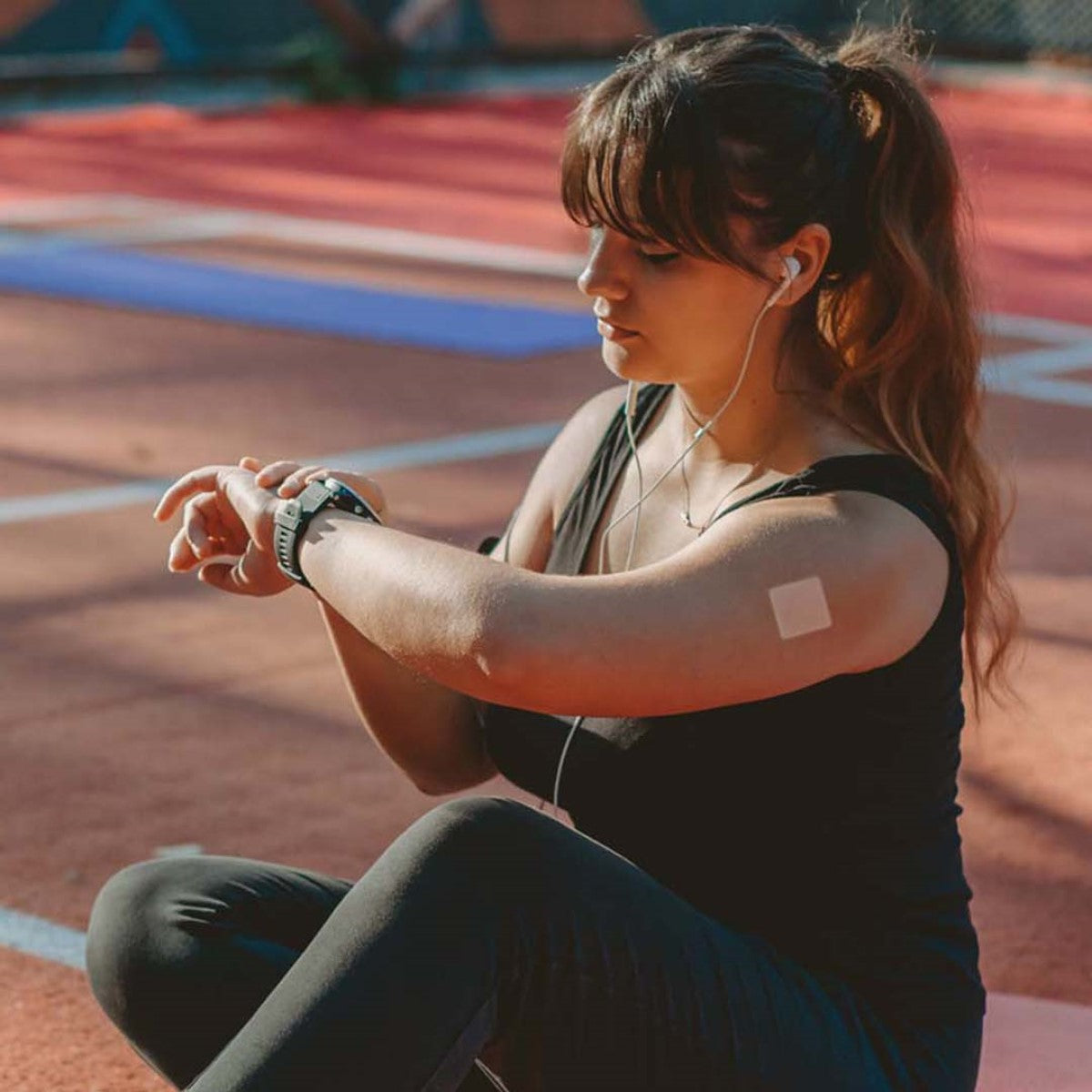 A woman wearing a black tank top and leggings sits on a sports court, checking her smartwatch while listening to music through earbuds, with a vitamin patch visible on her arm.