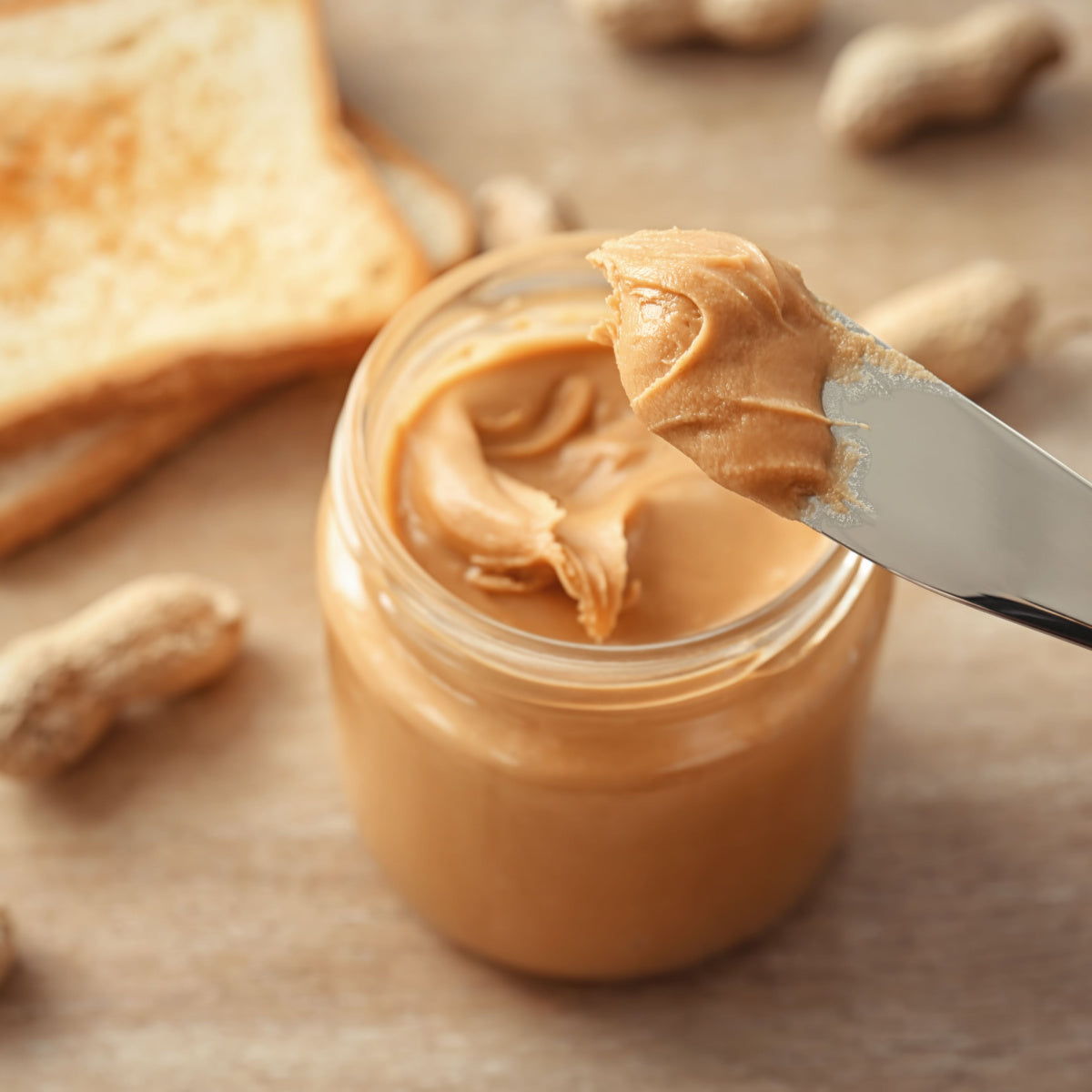 A knife holds a scoop of creamy peanut butter above a jar, with a slice of toasted bread and scattered peanuts in the background on a wooden surface.