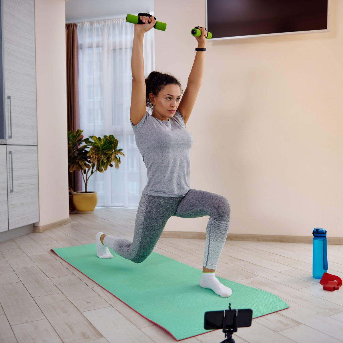 A woman in a gray workout outfit performs a lunge with green weights raised above her head on a yoga mat in a bright, modern living room with plants and exercise equipment nearby.