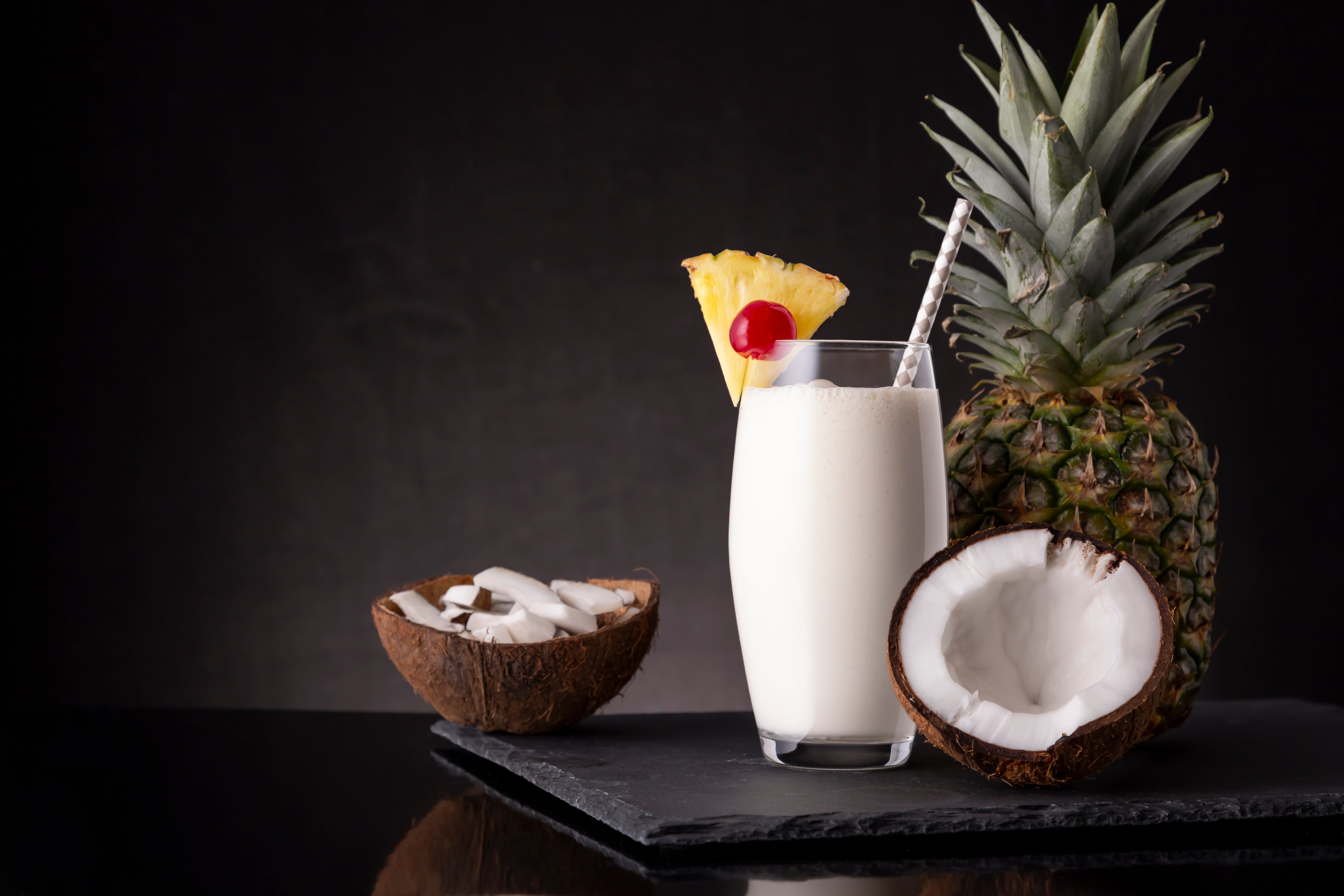 A creamy Pina Colada shake in a tall glass garnished with a pineapple slice and cherry sits on a slate board next to a whole pineapple and a halved coconut filled with coconut pieces.
