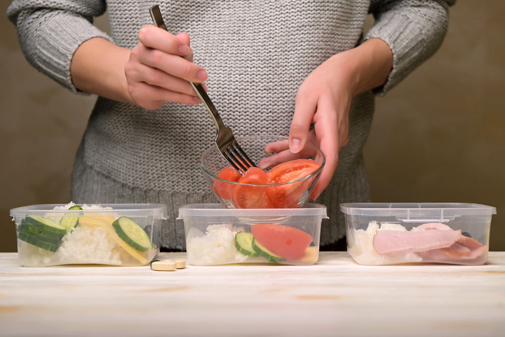 A person in a gray sweater uses a fork to transfer tomatoes from a bowl into three meal prep containers filled with rice, cucumbers, and sliced meat, emphasizing portion control for weight loss.