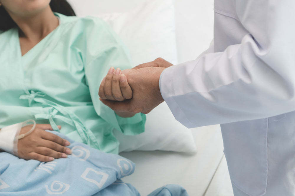 A patient in a hospital gown lies in bed while a doctor gently holds her hand, offering support and reassurance in a clinical setting.