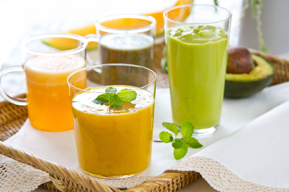A variety of colorful smoothies in clear glasses, including orange, green, and brown, are arranged on a woven tray, highlighting nutritious options for pre-op and post-operative bariatric diets.