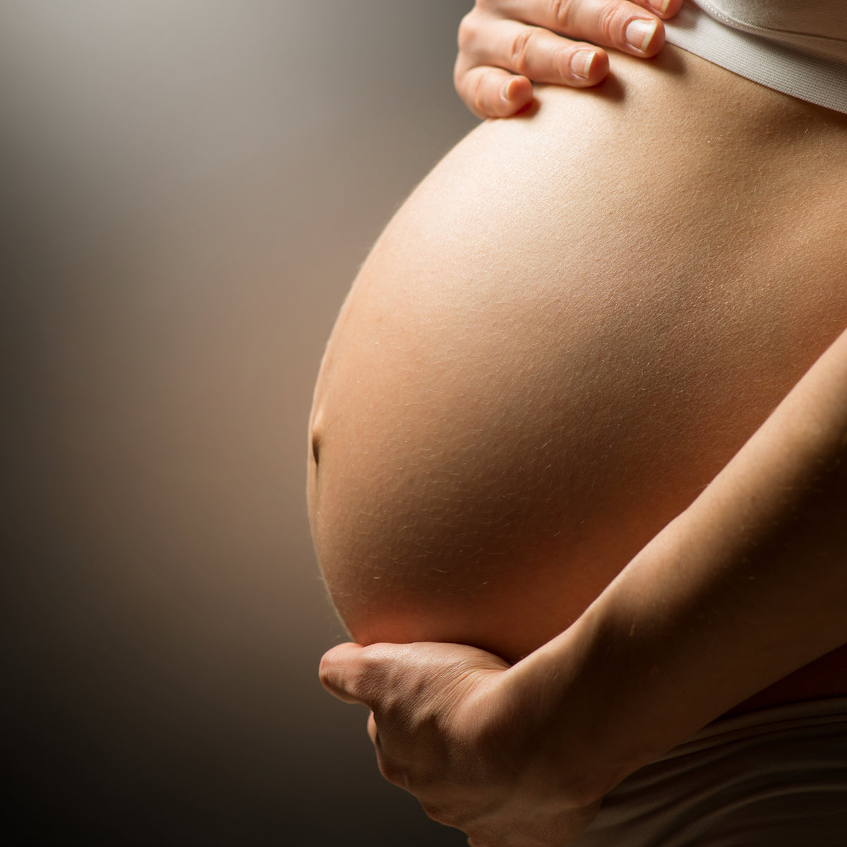 A close-up of a pregnant person's belly, gently cradled by one hand, set against a softly lit background, highlighting the pregnancy stage and the nurturing gesture.
