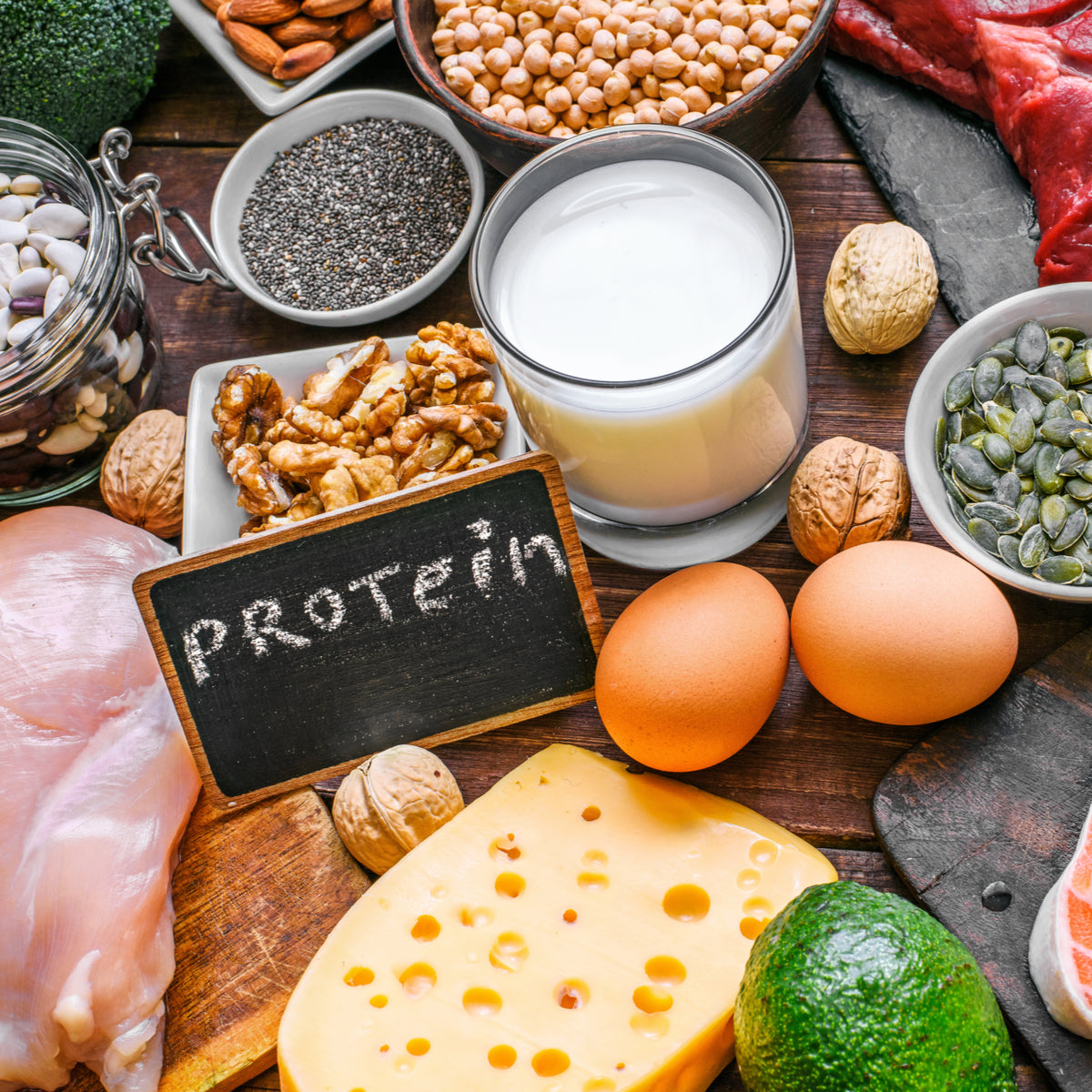 A variety of protein-rich foods, including chicken, eggs, cheese, nuts, seeds, and legumes, are arranged on a wooden surface with a small chalkboard displaying the word "protein."