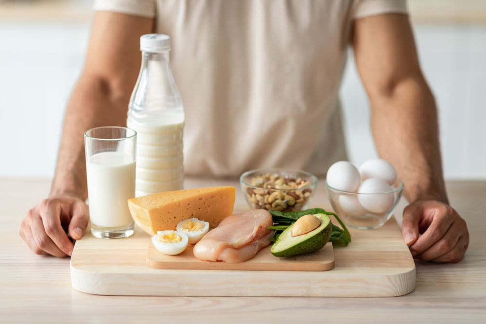A person in a light shirt presents a wooden board with various protein-rich foods, including chicken, eggs, cheese, milk, nuts, and avocado, emphasizing healthy eating for weight loss.