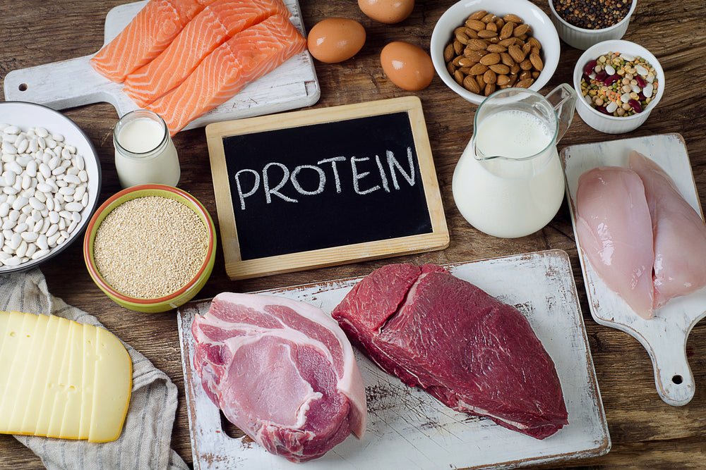 A variety of protein-rich foods, including salmon, chicken, eggs, beans, nuts, and dairy, are arranged on a wooden surface with a chalkboard displaying the word "PROTEIN."