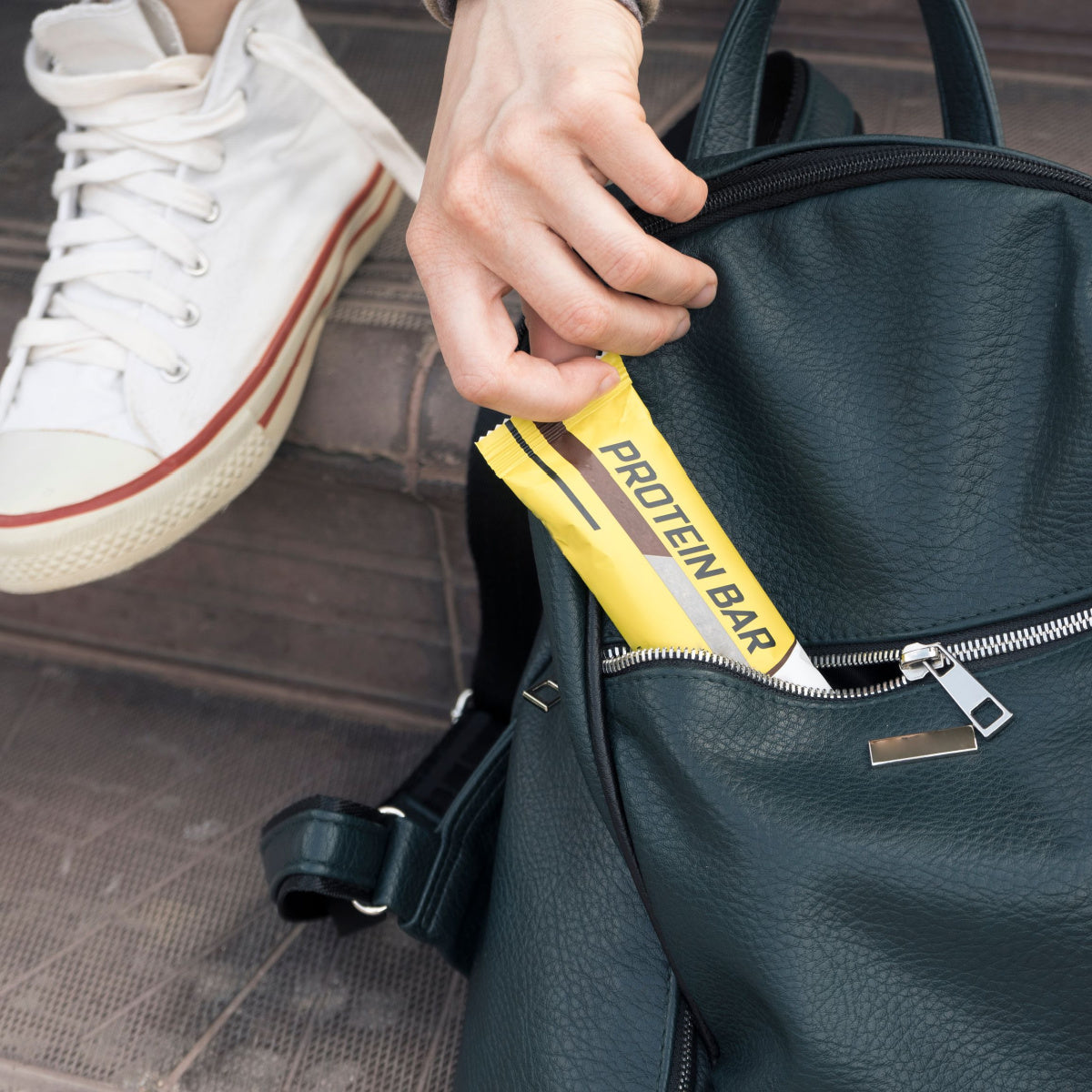 A hand reaches into a dark green backpack to pull out a yellow protein bar while a white sneaker rests on a step, suggesting a casual outdoor setting.