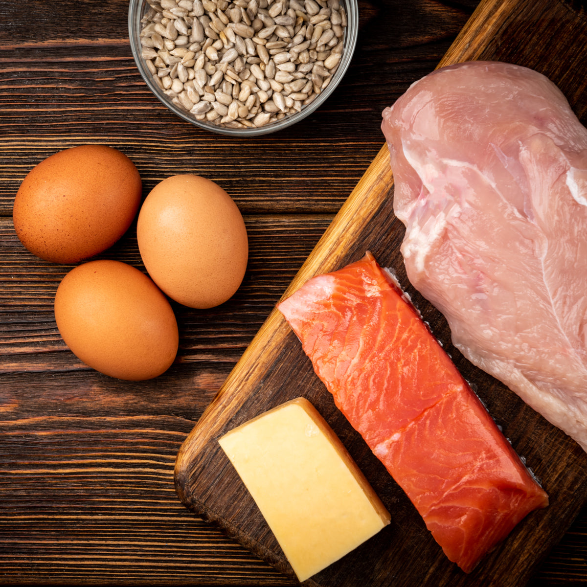 A wooden cutting board displays three brown eggs, a piece of chicken breast, a slice of salmon, and a block of cheese, with a bowl of sunflower seeds in the background, highlighting protein-rich foods for a diet plan.