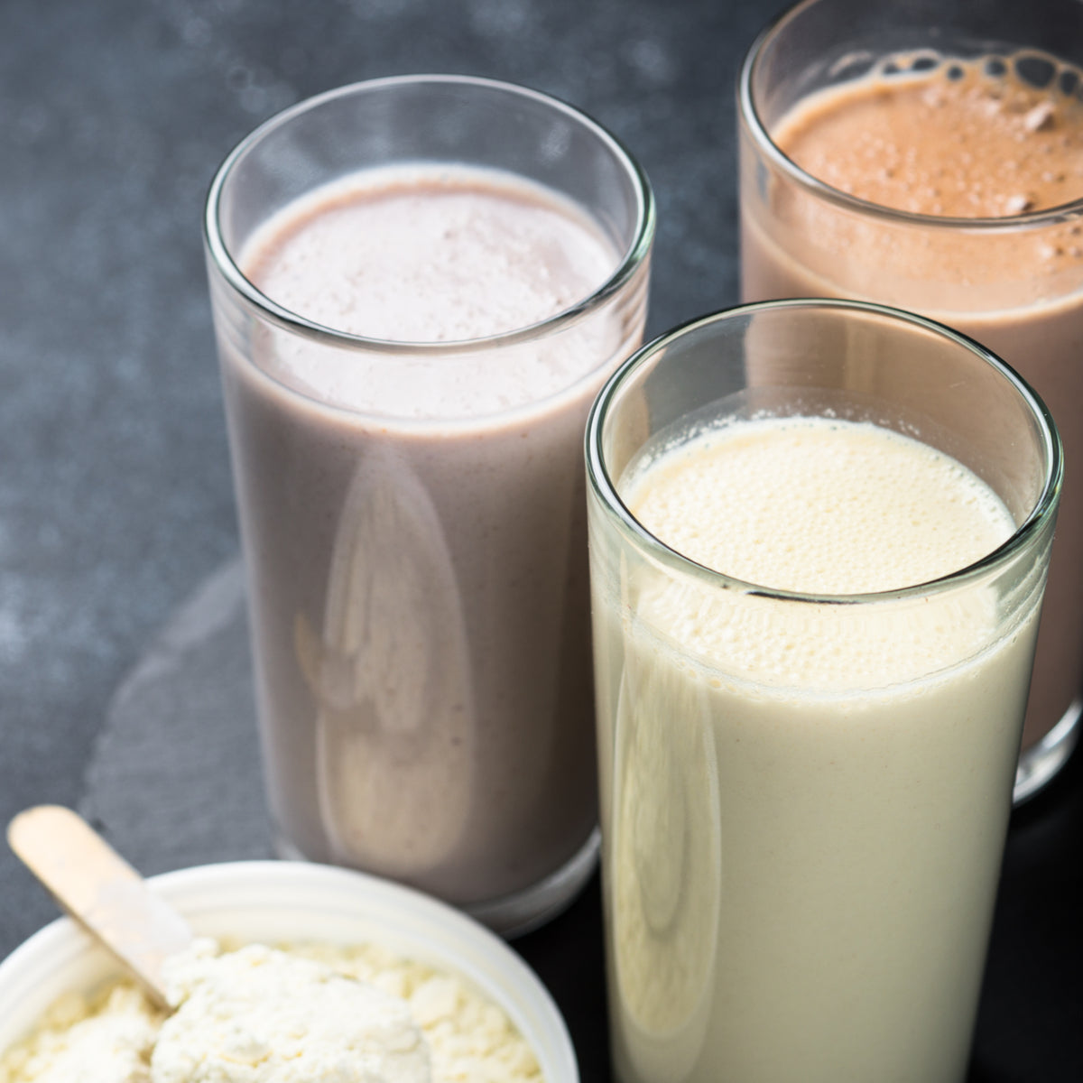 Three tall glasses of different protein shakes—light brown, dark brown, and creamy white—are arranged next to a bowl of protein powder with a scoop, set against a dark background.