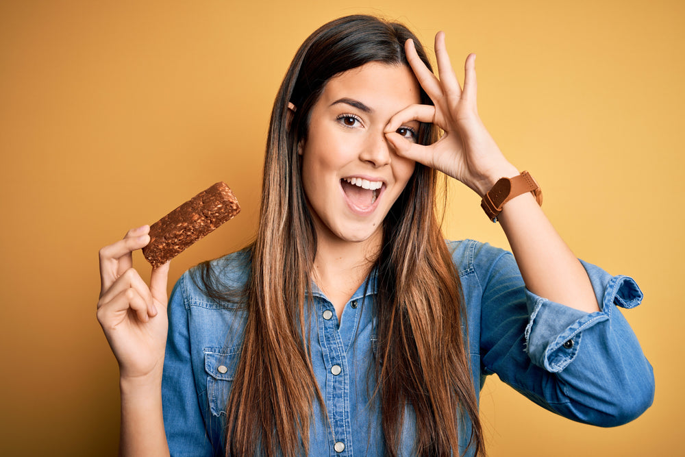 A young woman with long hair smiles and holds a protein bar in one hand while making an "OK" gesture with the other, set against a bright yellow background.