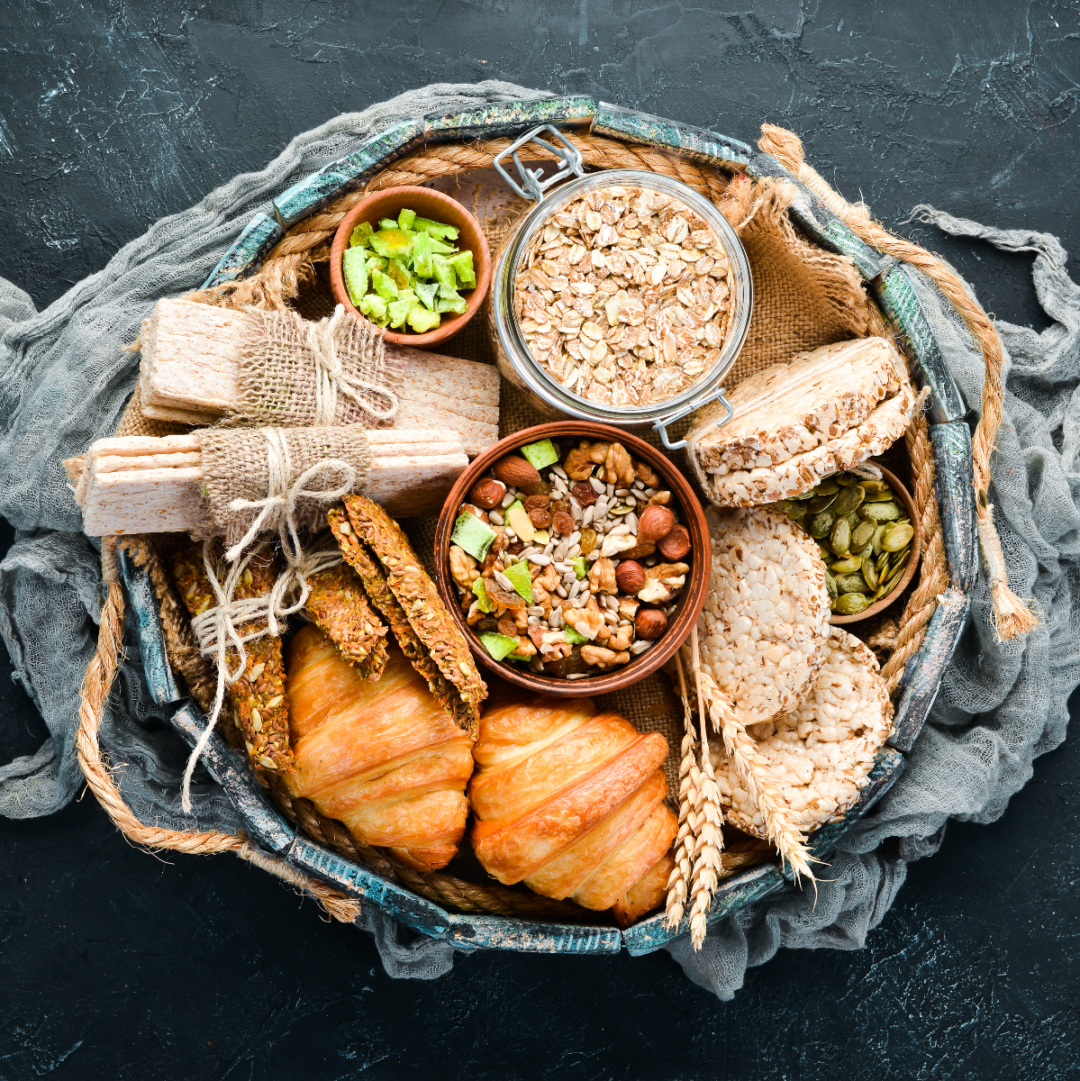 A woven basket filled with various protein-rich snacks, including croissants, granola, nuts, seeds, and rice cakes, arranged attractively on a textured dark background.