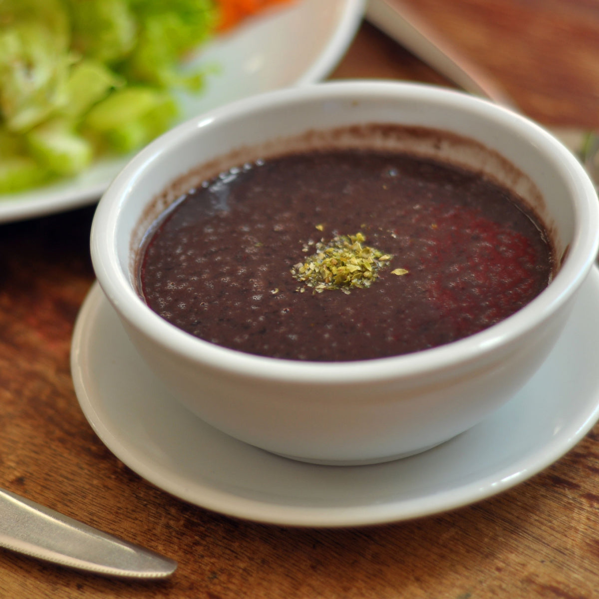 A white bowl filled with dark black bean soup, topped with a sprinkle of green herbs, sits on a wooden table, with a plate of fresh salad in the background.