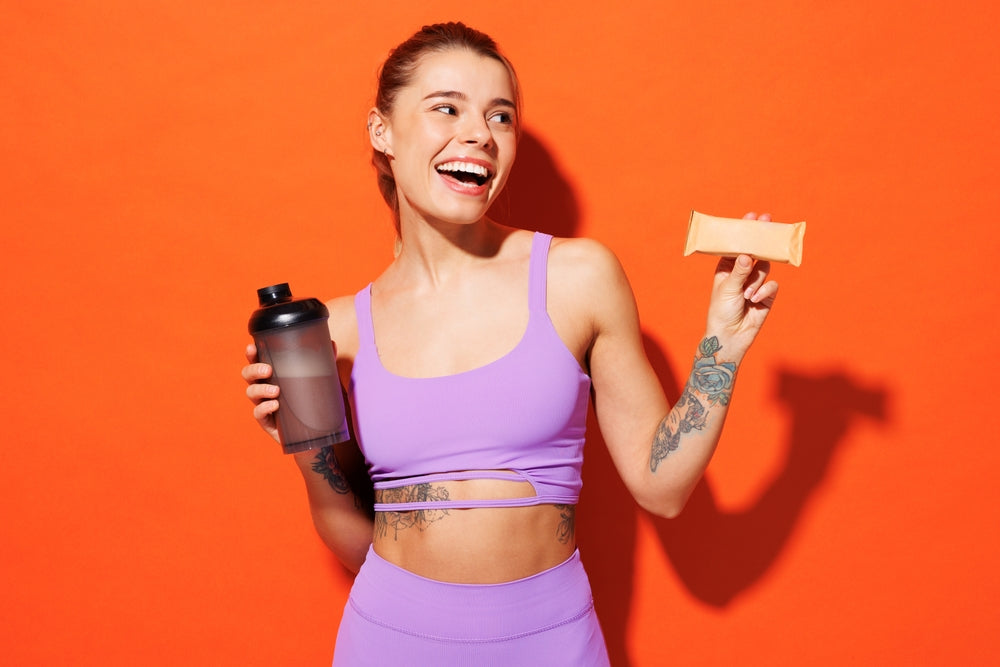 A smiling woman in a purple athletic outfit holds a protein shaker in one hand and a protein bar in the other against a bright orange background, promoting healthy nutrition.