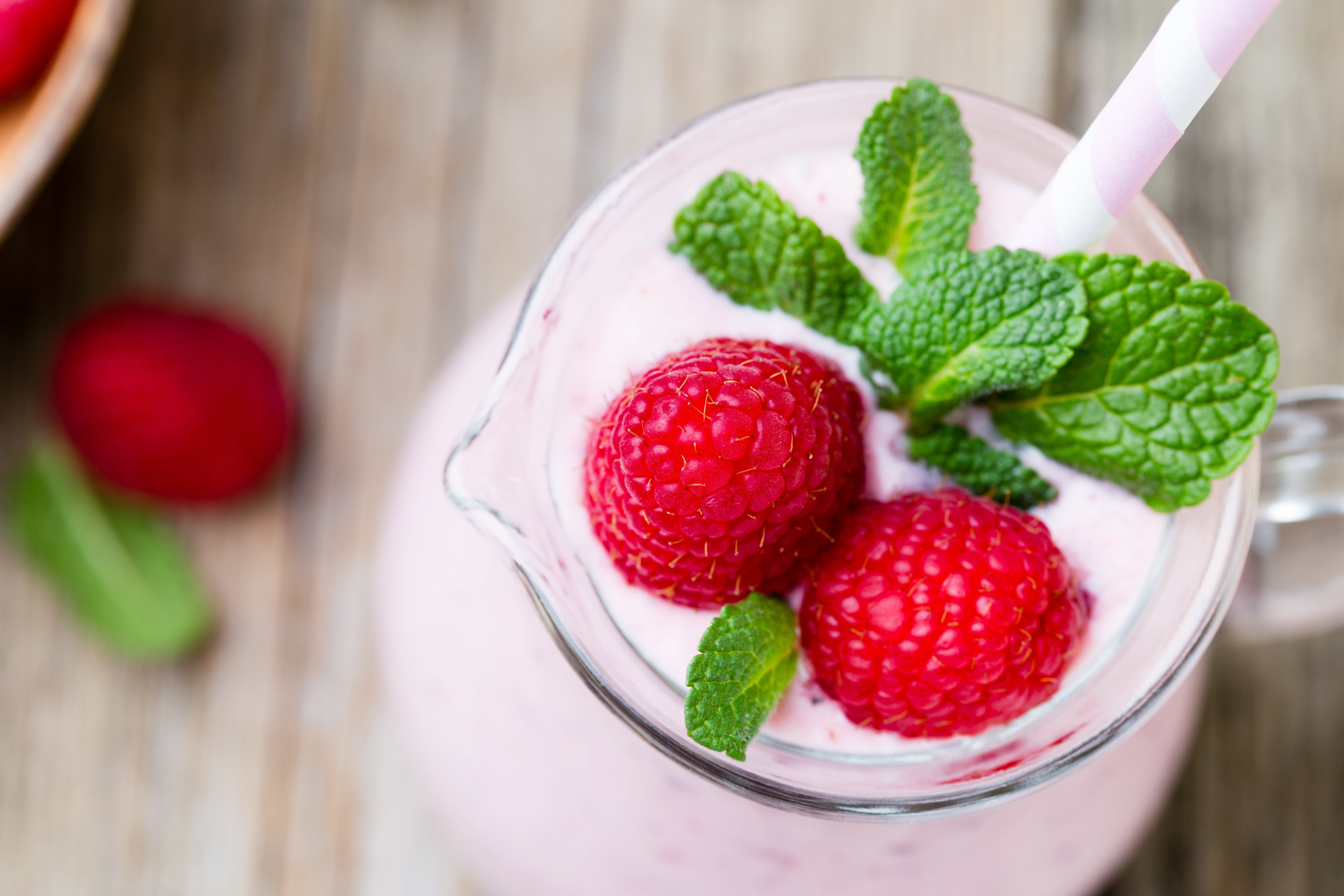 A glass of raspberry tea chiller shake topped with fresh raspberries and mint leaves, with a pink striped straw, is set on a wooden surface.
