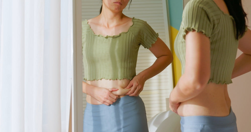 A woman in a green cropped top examines her midsection in a mirror, showing a thoughtful expression in a brightly lit room with colorful walls.