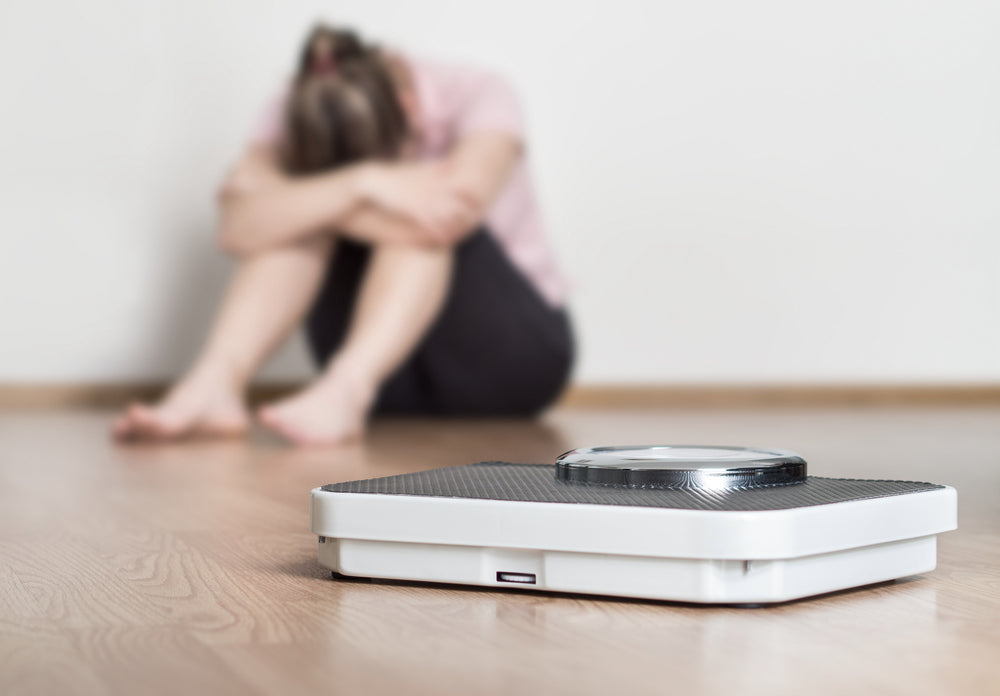 A white bathroom scale is in focus on the floor, while a person sits in the background, hunched over with their arms wrapped around their knees, conveying a sense of distress or disappointment.