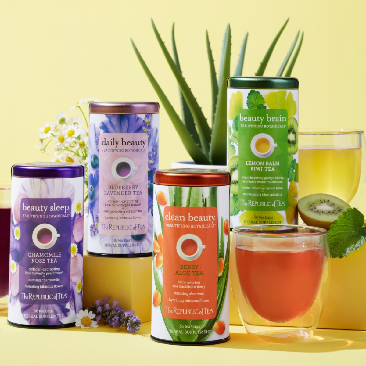 Five colorful tea canisters from The Republic of Tea are displayed against a yellow background, alongside two glasses of tea and an aloe plant, highlighting various herbal blends for beauty and wellness.