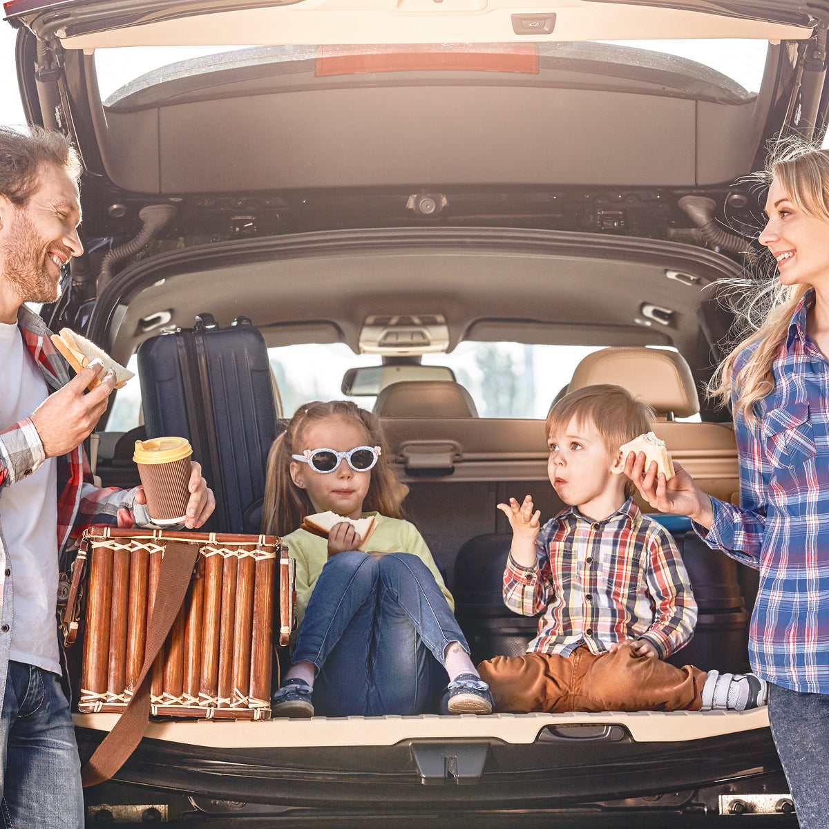 A smiling family enjoys road trip snacks in the back of a car, with a father holding a sandwich and coffee, while two children sit on the cargo area, one wearing sunglasses and eating a sandwich.