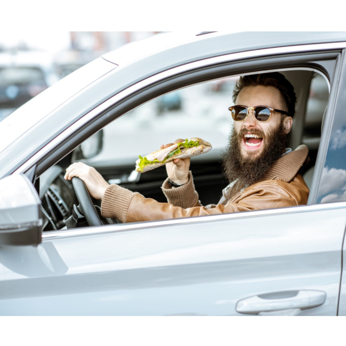 A man with a beard and sunglasses smiles widely while holding a sandwich in his right hand, sitting in the driver's seat of a car parked in a lot.