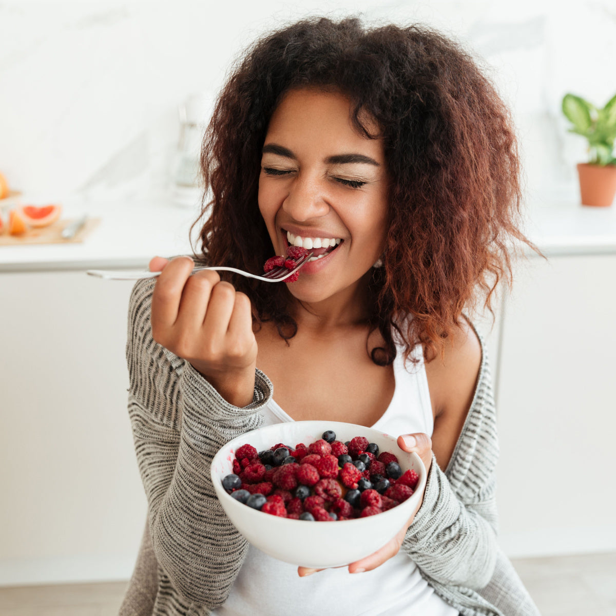 A woman with curly hair smiles joyfully while holding a bowl of mixed berries and taking a bite with a fork, seated in a bright kitchen with plants in the background.