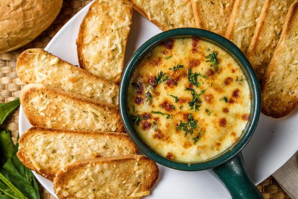 A bowl of melted cheese dip topped with herbs sits in the center, surrounded by crispy, toasted bread slices on a white plate, with a woven mat in the background.