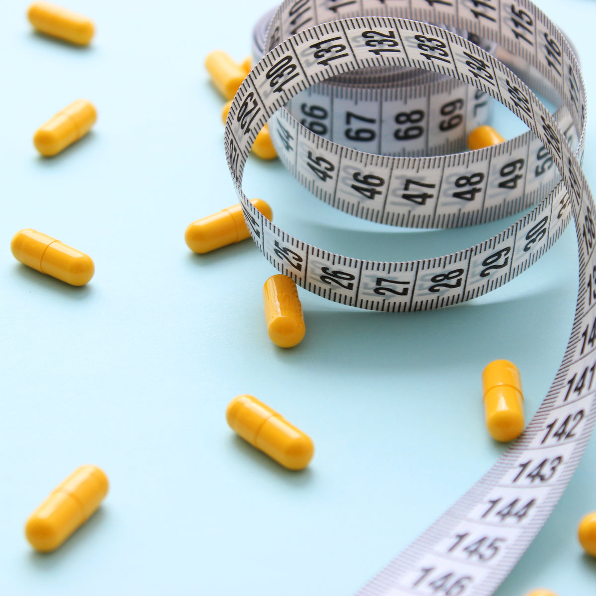 Yellow capsules are scattered on a light blue surface, with a measuring tape coiled around some of the capsules, suggesting a focus on weight loss and fat burning supplements.