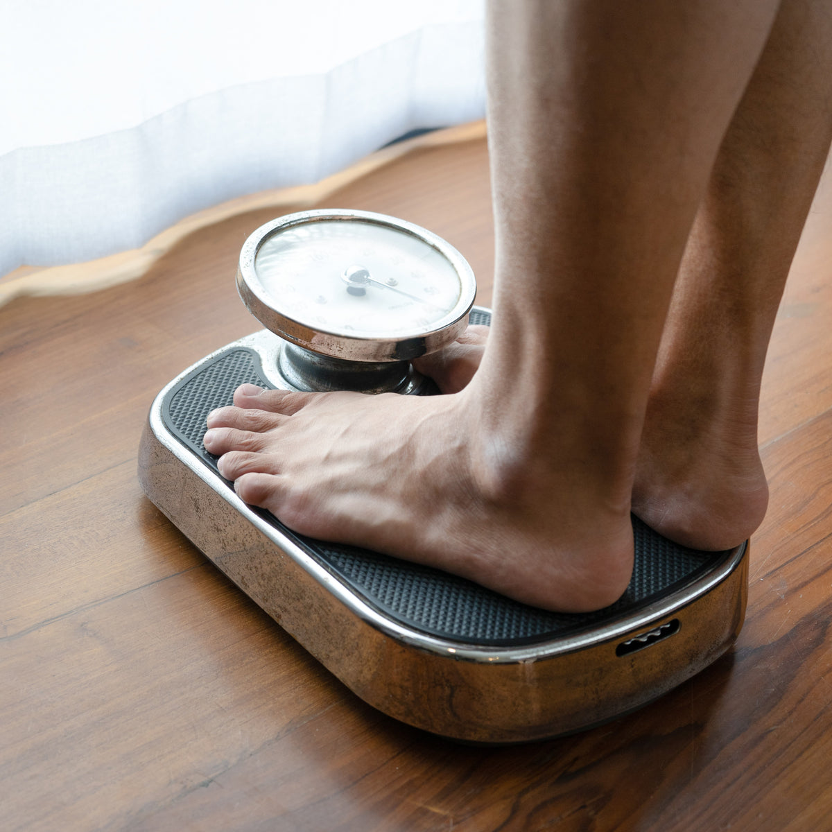 A person stands on a vintage bathroom scale with bare feet, measuring their weight on a wooden floor, indicating a focus on weight management or health monitoring.